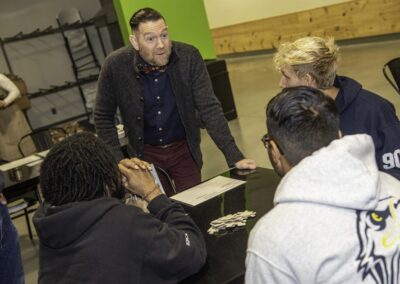 A man in a grey sweater stands at a table while 3 other people sit at the table facing him