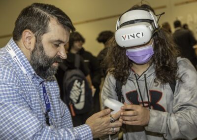 Student with a VR headset on stands next to a man who is handing him VR controlers