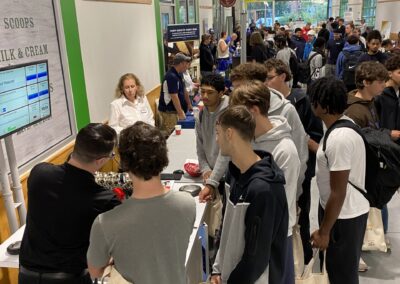 High School students gather around exhibitor tables in a long hallway.