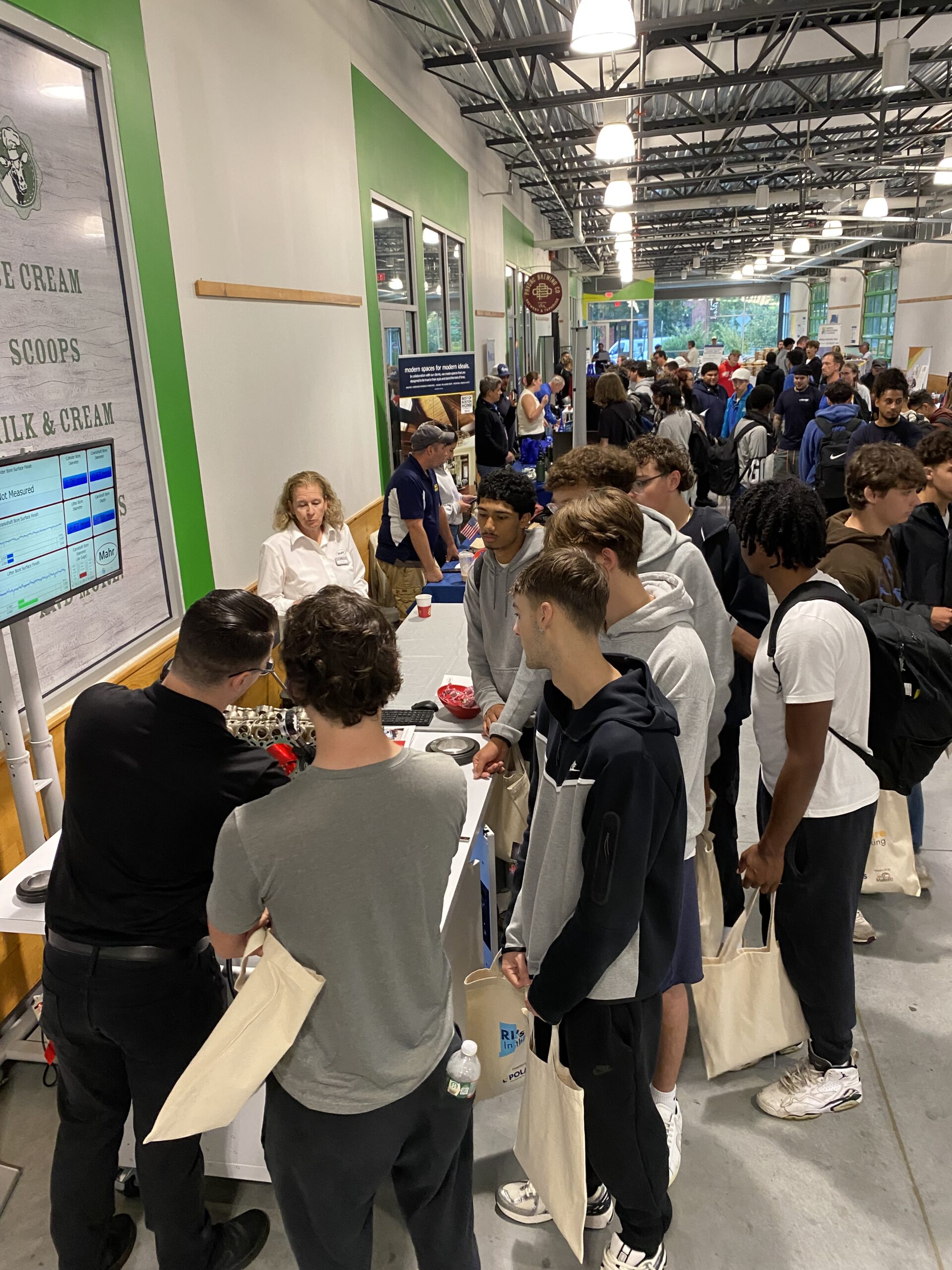 High School students gather around exhibitor tables in a long hallway.